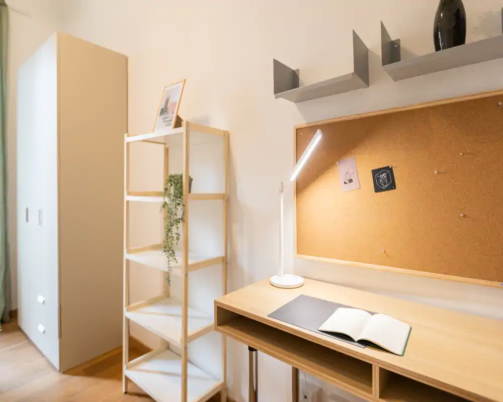 Angolo camera luminoso e minimal con scrivania in legno e lampada, bacheca in sughero, scaffalatura aperta e armadio — ideale per studio o smart working.
