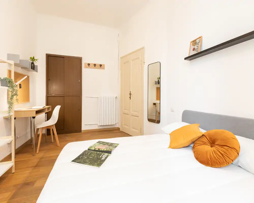 Camera da letto luminosa e ben arredata con letto bianco, cuscini arancioni caldi e una scrivania nell'angolo — ideale per affitti brevi o studenti.