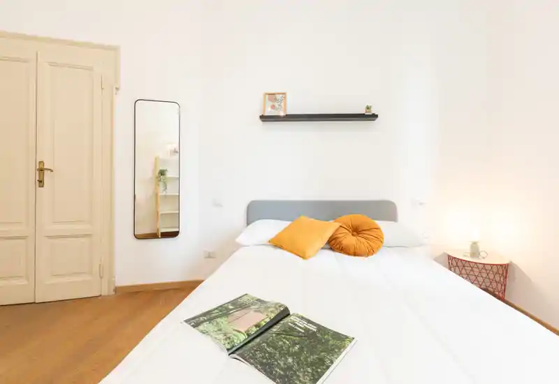 Camera da letto luminosa e minimal con un letto ben rifatto, cuscini colorati, lampada da comodino e una mensola semplice: uno spazio moderno e tranquillo perfetto per riposare.