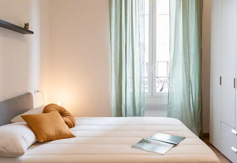 Camera da letto luminosa e accogliente con letto matrimoniale ben rifatto, biancheria morbida, cuscini decorativi e luce naturale da una finestra con tende verde chiaro.