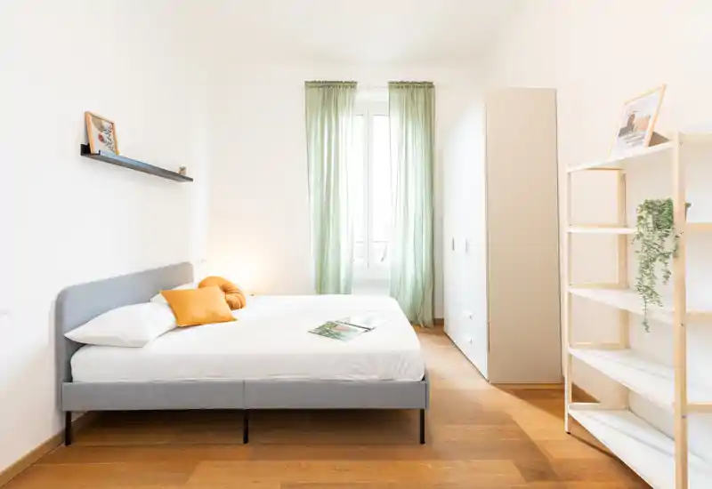 Camera da letto luminosa e minimal con letto matrimoniale, struttura imbottita grigia, pavimento in legno caldo e tende leggere verdi che filtrano la luce naturale.