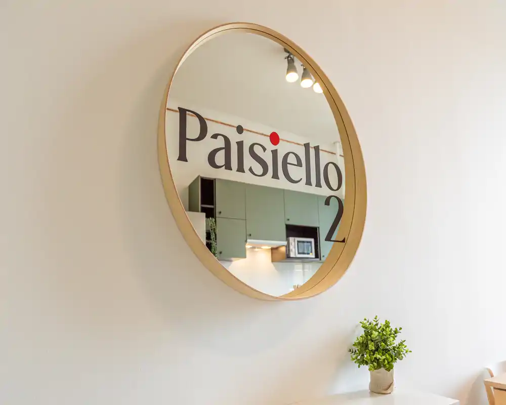 Un grande specchio rotondo che riflette una cucina moderna con pensili verdi, forno a microonde e luci sotto i pensili; una piccola pianta in vaso è appoggiata vicino.