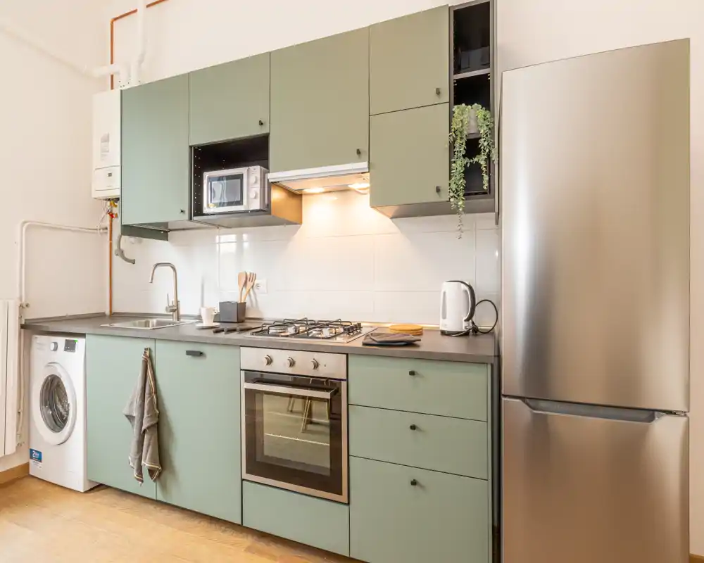 Cucina compatta e moderna con mobili verdi, elettrodomestici in acciaio, piano cottura a gas e forno integrato. Stile pulito e buona luce naturale e artificiale, perfetta per annunci.