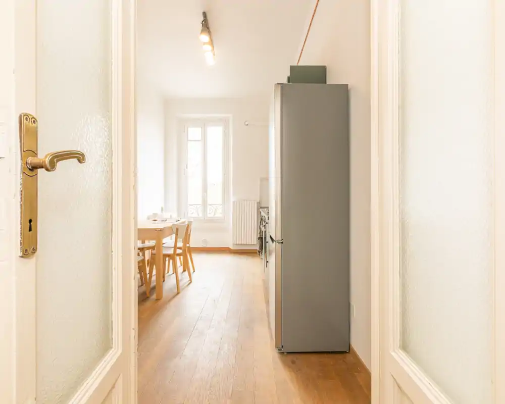 Cucina luminosa e minimal con pavimento in legno, piccolo tavolo da pranzo e sedie accanto a un frigorifero alto; spazio pulito e ben illuminato ideale per appartamenti compatti.
