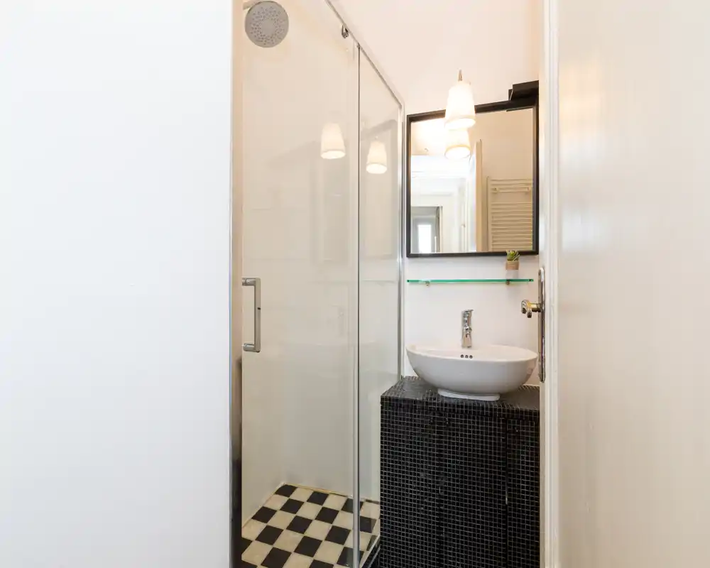Bagno compatto e moderno con box doccia in vetro, lavabo a ciotola bianco su mobile con piastrelle nere e specchio con applique. Spazio luminoso, pulito e adatto per annunci di affitto.