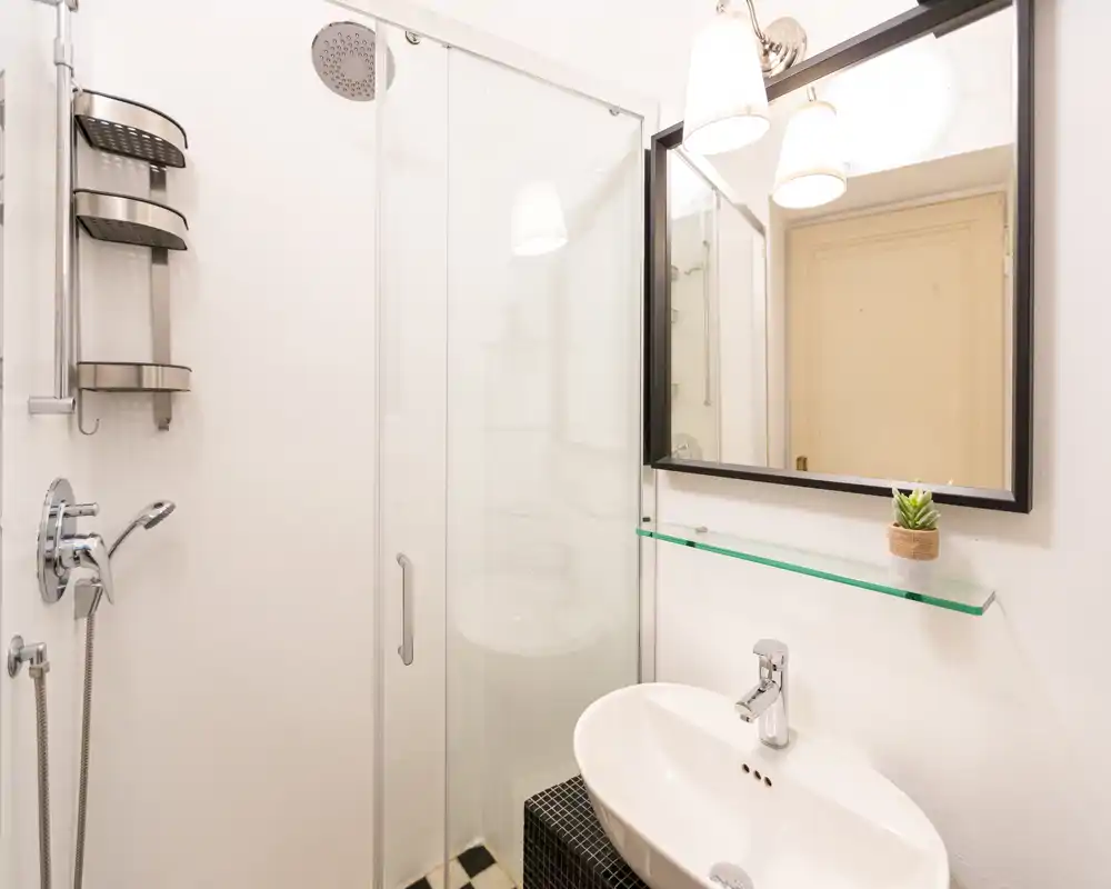 Bagno moderno e luminoso con box doccia in vetro, rubinetteria a muro e lavabo bianco sotto uno specchio incorniciato.