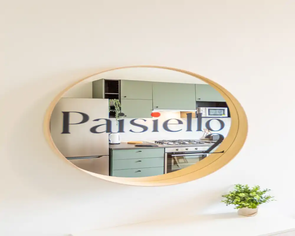 Cucina moderna vista attraverso uno specchio circolare: mobili verdi, frigorifero in acciaio, piano cottura a gas e forno visibili, con una piccola pianta che aggiunge freschezza.