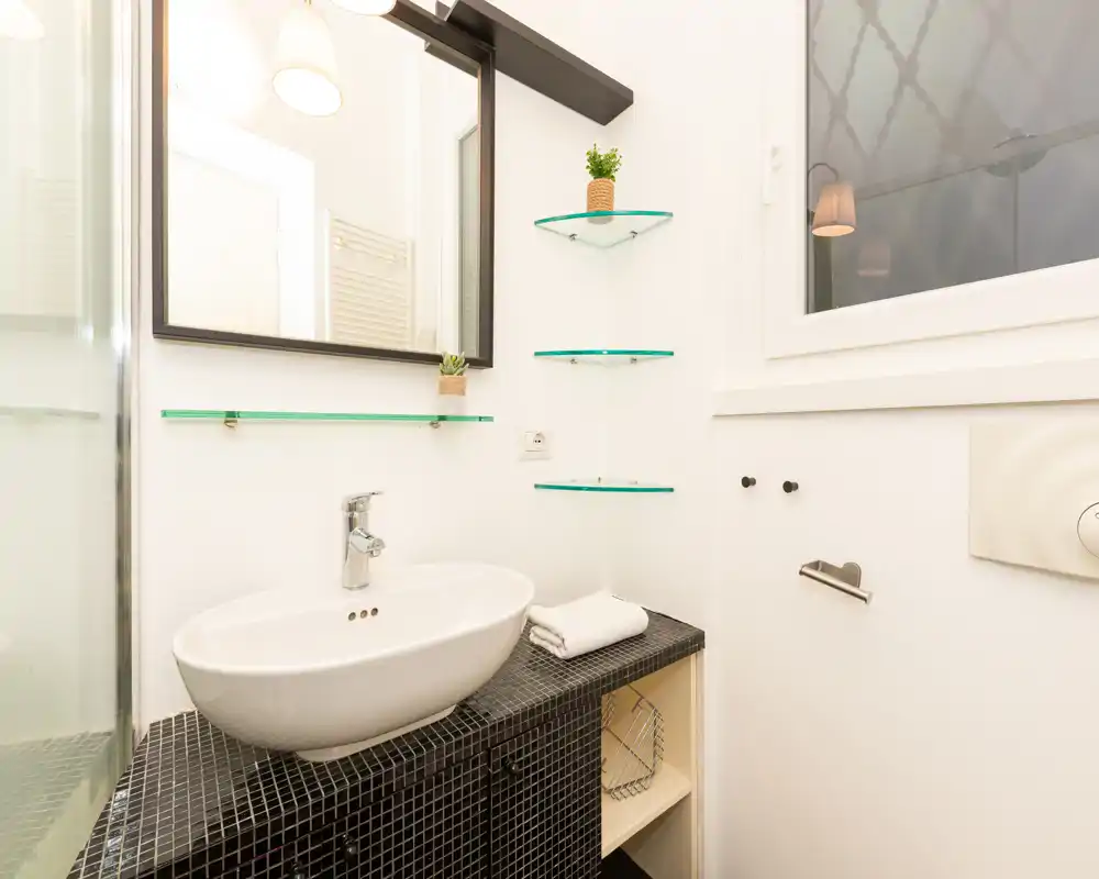 Bagno luminoso e moderno con lavabo a bacinella su mobile piastrellato, specchio a parete e mensole in vetro accanto a una finestra opaca.