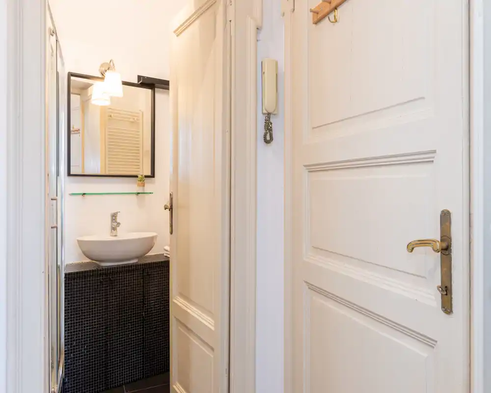 Bagno compatto fotografato attraverso la porta aperta: lavabo a ciotola moderno, mobile con piastrelle mosaico, specchio e applique a parete — luminoso e ordinato.