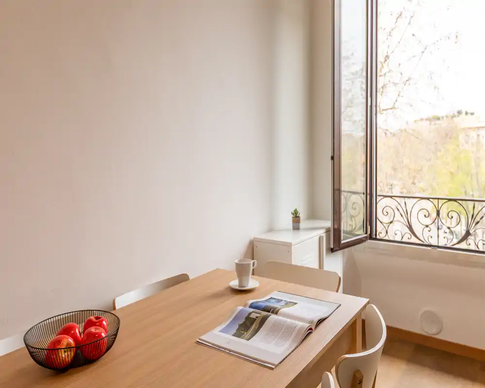 Zona pranzo illuminata dal sole con un tavolo in legno, sedie, una ciotola di mele e una finestra aperta che porta luce naturale — semplice, calda e accogliente per pasti o lavoro.