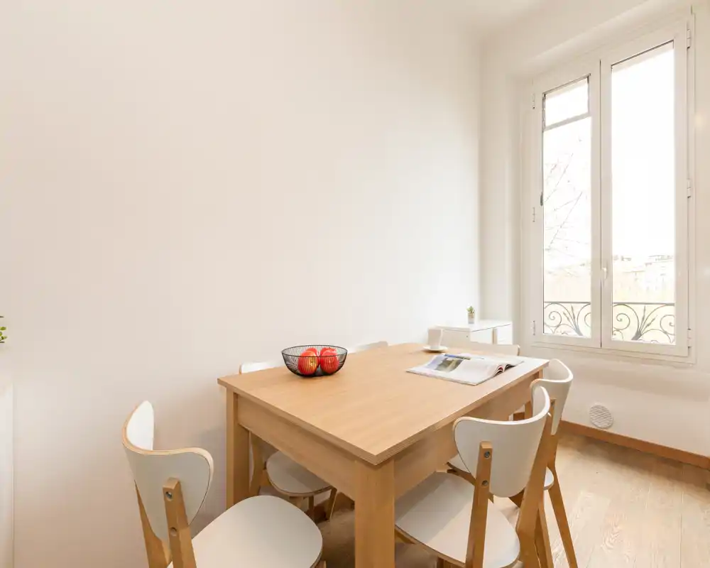 Zona pranzo luminosa e minimal con tavolo in legno e quattro sedie accanto a una grande finestra che lascia entrare luce naturale.