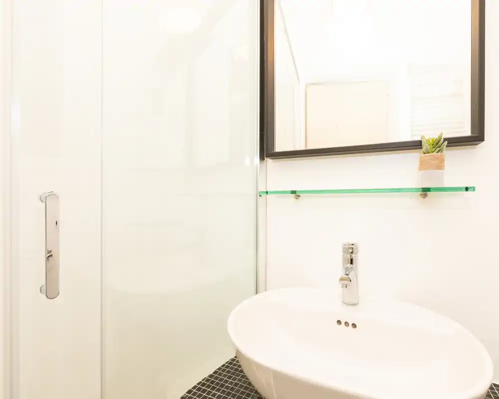 Bagno moderno e luminoso con lavabo bianco, rubinetto cromato, porta doccia in vetro e specchio sopra una sottile mensola in vetro con una piccola pianta.