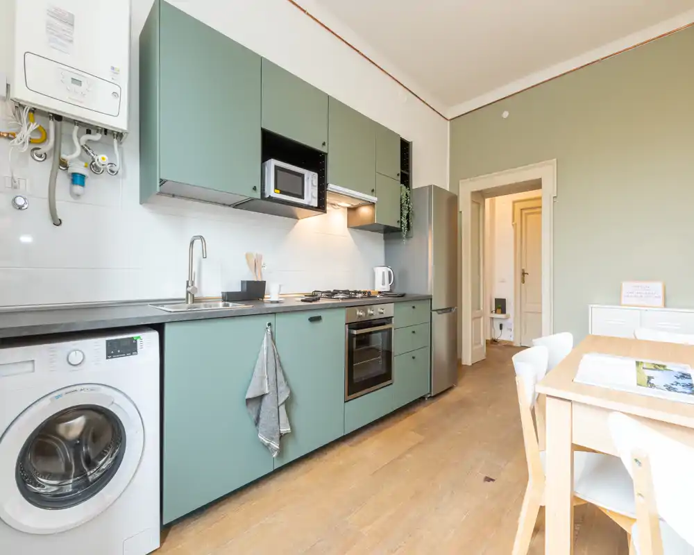 Cucina moderna con mobili verde salvia, forno e frigorifero integrati, lavatrice e un tavolo in legno — luminosa, ordinata e adatta a affitti brevi.