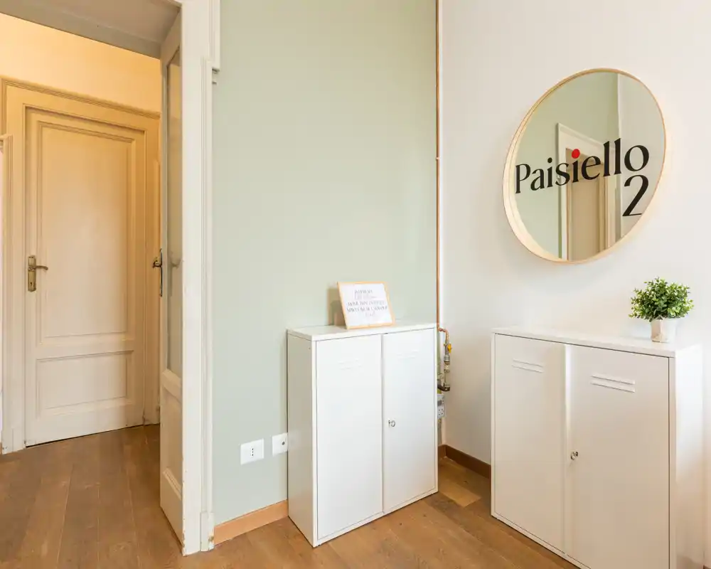 Ingresso luminoso e ordinato con pavimento in legno, mobili contenitori bianchi e uno specchio rotondo decorativo che riflette l’interno.