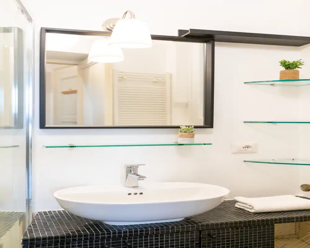 Bagno moderno e luminoso con lavabo elegante, grande specchio, mensole in vetro e piano piastrellato — pulito e ben illuminato.