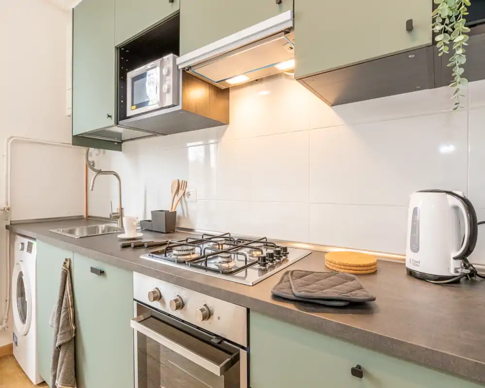 Cucina compatta e moderna con ante verde menta, piano cottura a gas, forno incassato, microonde e piccoli elettrodomestici; luminosa, ordinata e pronta all'uso.