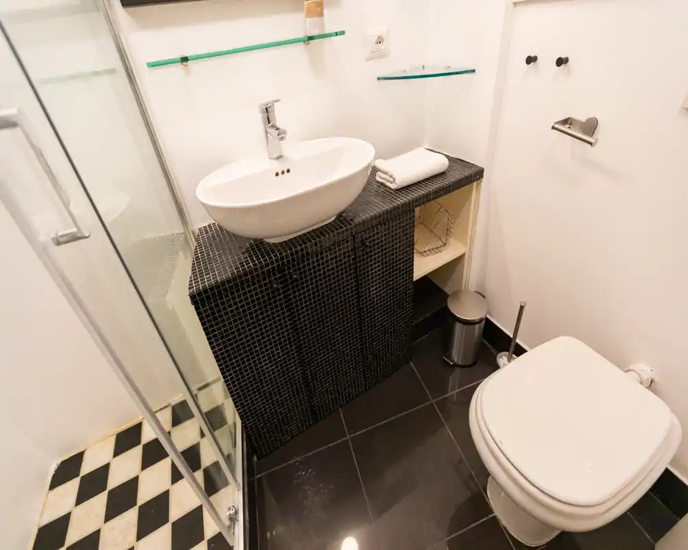 Bagno moderno e compatto con lavabo a bacinella su mobile rivestito in mosaico nero, box doccia in vetro e wc a parete — pavimenti piastrellati e mensole in vetro semplici.