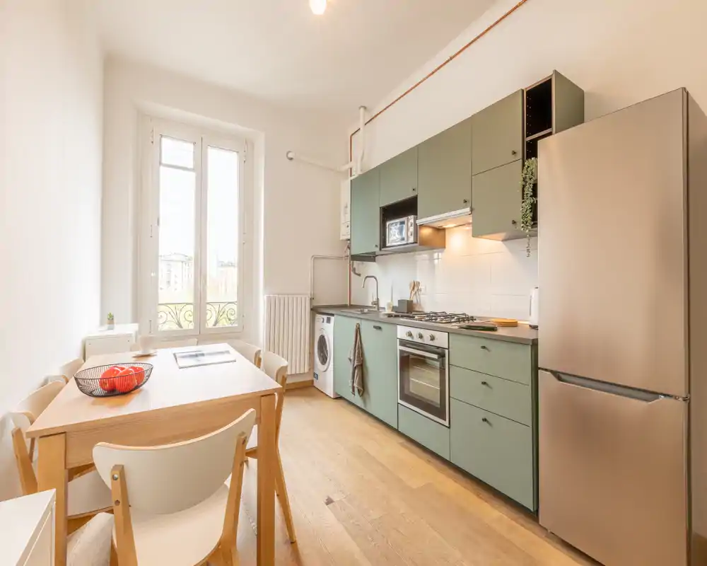 Cucina luminosa e moderna con mobili verde salvia, forno integrato e frigorifero, lavatrice e un accogliente tavolo in legno vicino a una grande finestra.