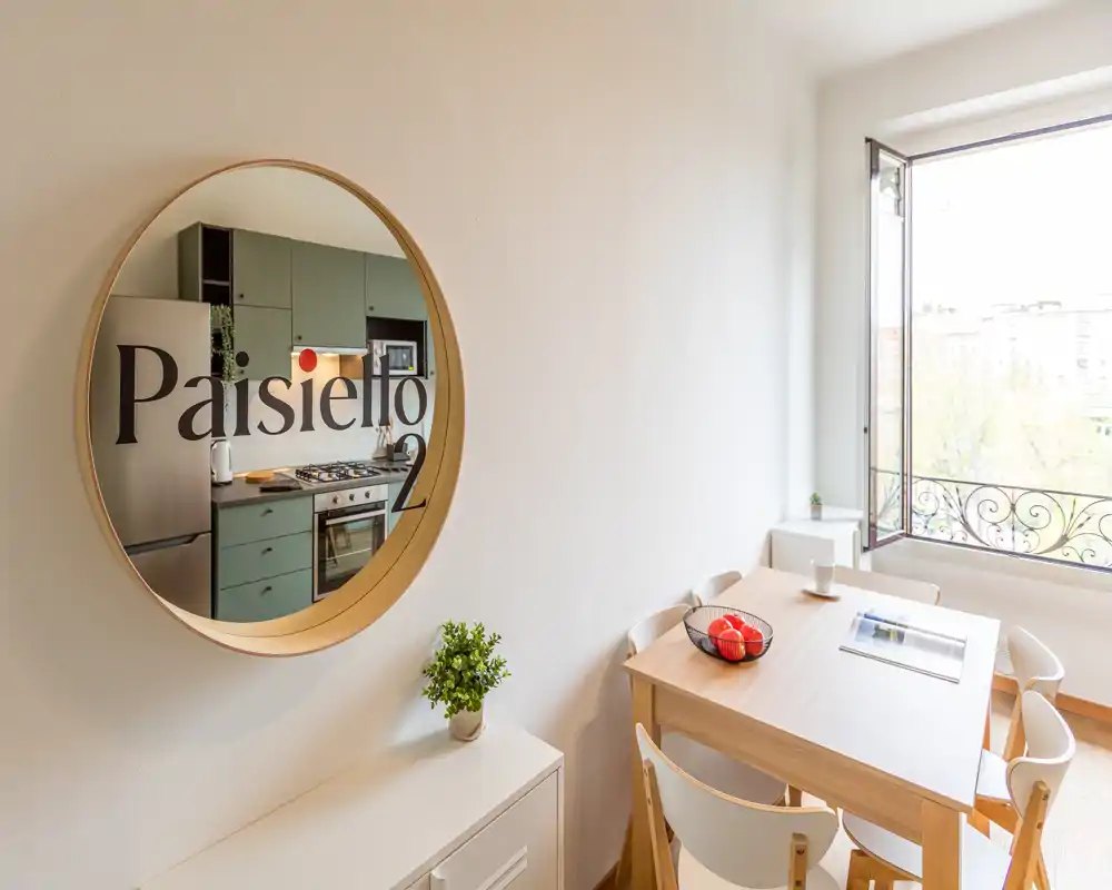 Angolo pranzo luminoso con tavolo in legno per quattro, sedie bianche e finestra aperta; uno specchio rotondo riflette la cucina adiacente aggiungendo profondità e stile.