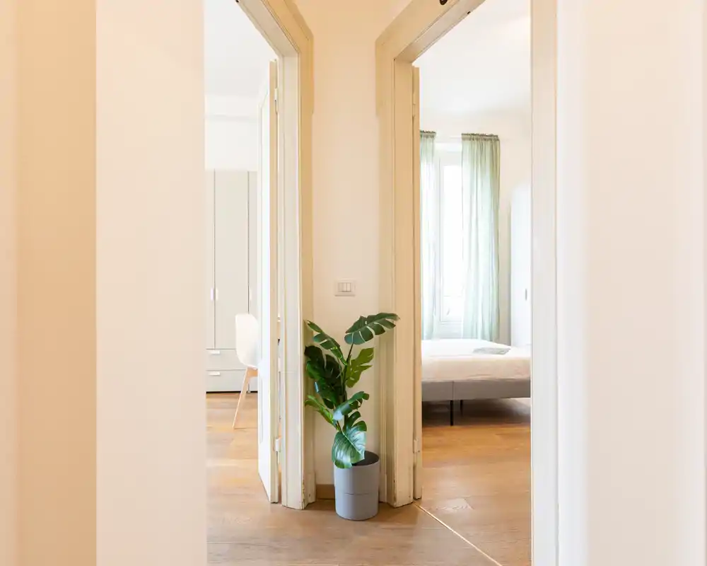 Camera da letto luminosa e minimal vista attraverso l’ingresso del corridoio, con letto vicino alla finestra, tende chiare e pavimenti in legno che creano un’atmosfera ariosa.