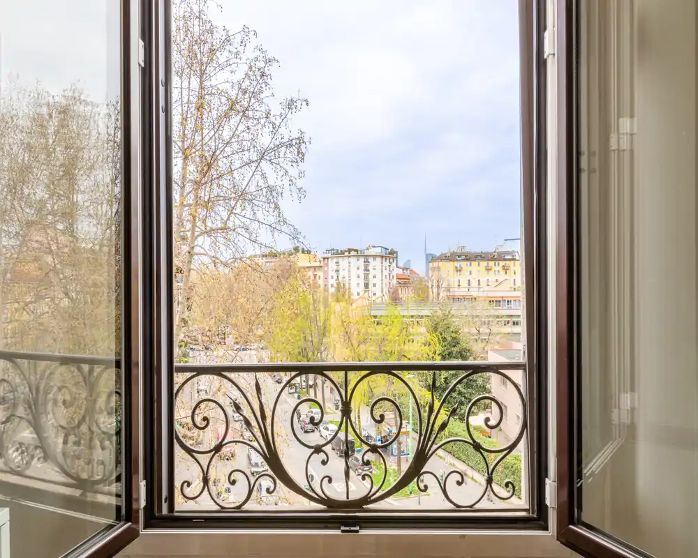 Finestra aperta con ringhiera in ferro battuto decorativa che si affaccia su una strada alberata e sugli edifici della città — vista urbana luminosa e libera.