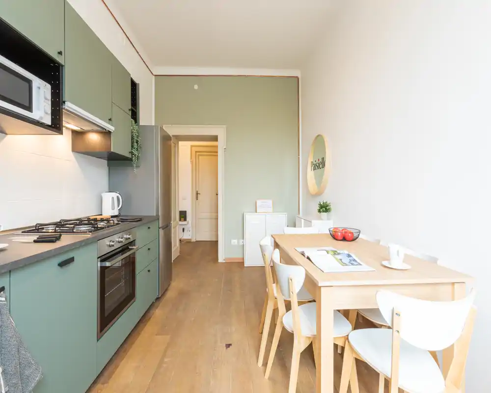 Angolo cottura moderno con elettrodomestici integrati e tavolo da pranzo in legno chiaro per quattro persone. Stile luminoso e fresco con pensili verdi e linee pulite, perfetto per affitti brevi.