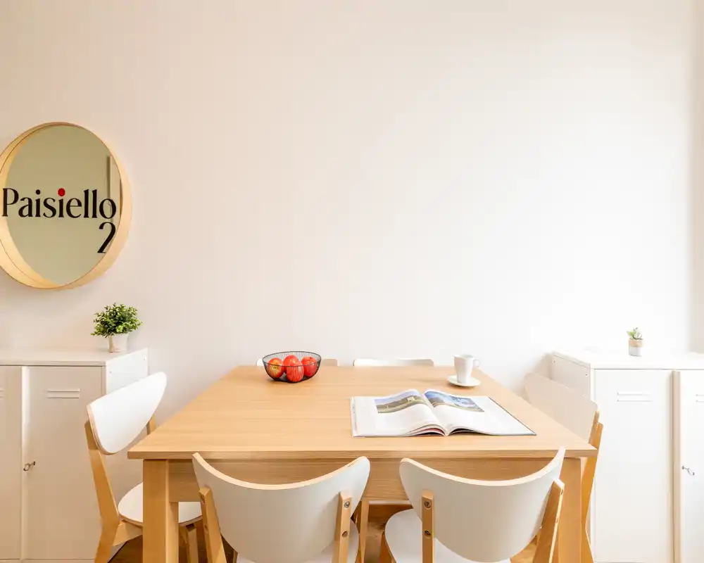 Zona pranzo luminosa e minimal con tavolo in legno, quattro sedie moderne, ciotola decorativa con frutta e una piccola pianta — ideale per pasti accoglienti o smart working.