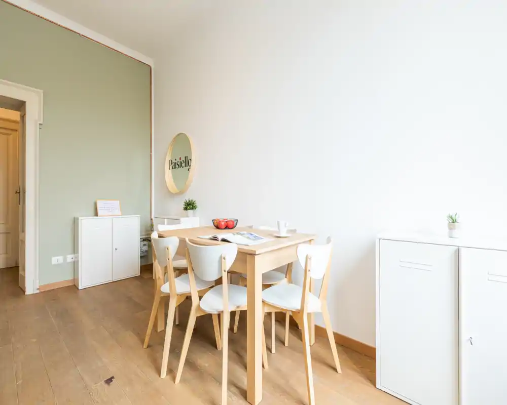 Zona pranzo luminosa e minimal con tavolo in legno chiaro e quattro sedie bianche, armadietti semplici e decorazioni discrete, perfetta per un appartamento compatto.