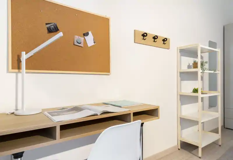 Angolo studio luminoso e minimal all'interno di una zona giorno: scrivania in legno chiaro con lampada, pannello in sughero, scaffale aperto e sedia semplice — pulito, funzionale e ben illuminato.