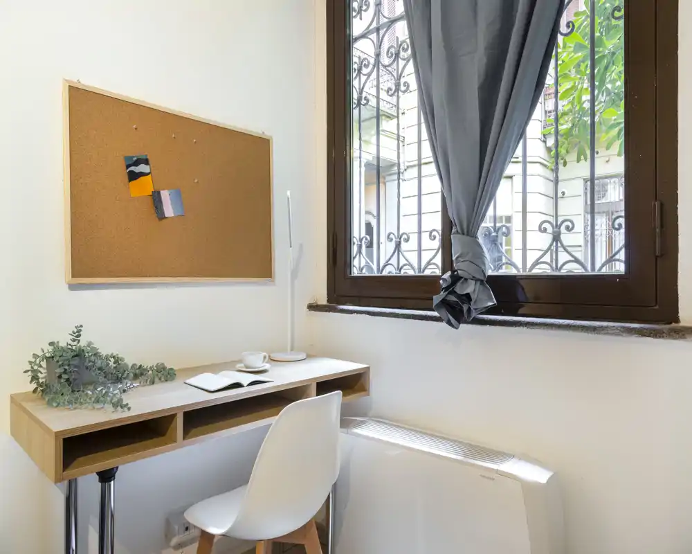 Angolo studio compatto con scrivania in legno, sedia e una bacheca in sughero accanto a una finestra che lascia entrare luce naturale. Piccoli dettagli decorativi come una pianta e una tazza rendono lo spazio ordinato e funzionale.