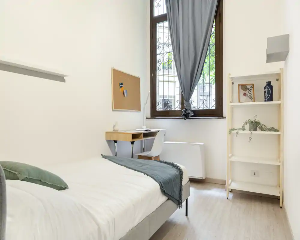 Camera singola luminosa e minimal con letto ben rifatto, scrittoio accanto alla finestra e scaffale semplice — ideale per studenti o una persona sola.