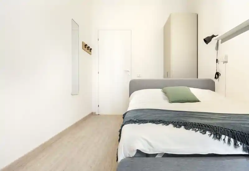 Bright, minimal double bedroom with a grey upholstered bed, green cushion and a textured throw; neutral walls, wardrobe and a bedside lamp create a calm, modern feel.