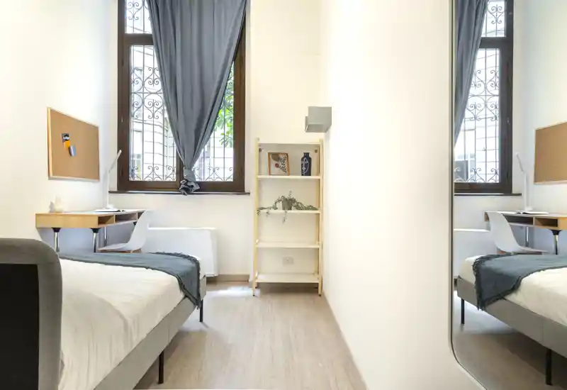 Bright, minimalist bedroom with a neatly made bed, small study desk by the window and a slim shelving unit; natural light and clean lines create a calm atmosphere.