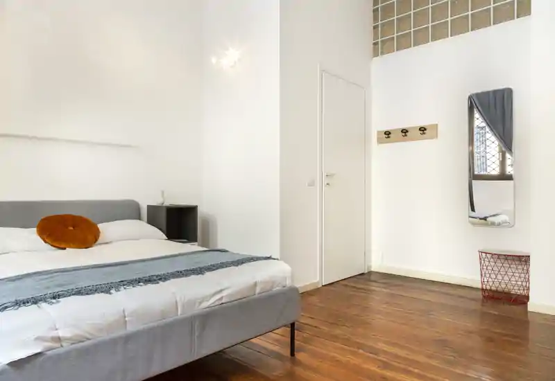 Camera da letto moderna e luminosa con letto imbottito grigio, pavimento in legno caldo e arredi semplici e curati, con cuscino rotondo arancione e specchio a parete.