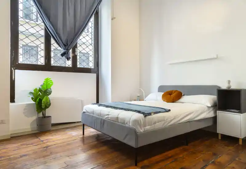Camera da letto moderna e luminosa con letto matrimoniale ben rifatto, pavimenti in legno caldi e una grande finestra che illumina l’ambiente.