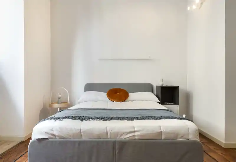 Camera da letto luminosa e minimalista con letto matrimoniale posizionato al centro, testiera grigia, biancheria bianca e un cuscino decorativo arancione. Linee pulite, pavimento in legno e comodini semplici creano un ambiente tranquillo e accogliente, perfetto per annunci.