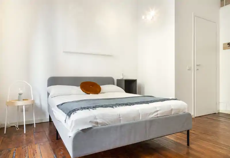 Camera da letto luminosa e moderna con un letto matrimoniale completamente visibile, biancheria morbida, un cuscino decorativo rotondo e pavimenti in legno che creano un'atmosfera calda e minimal.