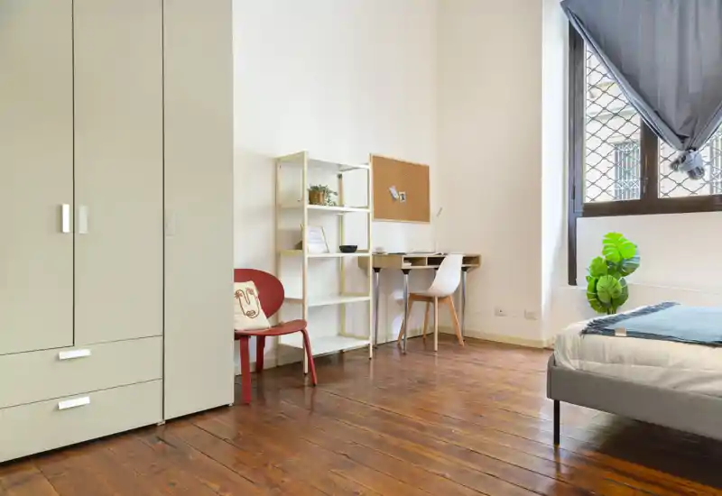 Camera da letto luminosa e minimal con letto visibile, pavimento in legno, armadio e una scrivania vicino alla finestra, ideale per una persona o uno studente.