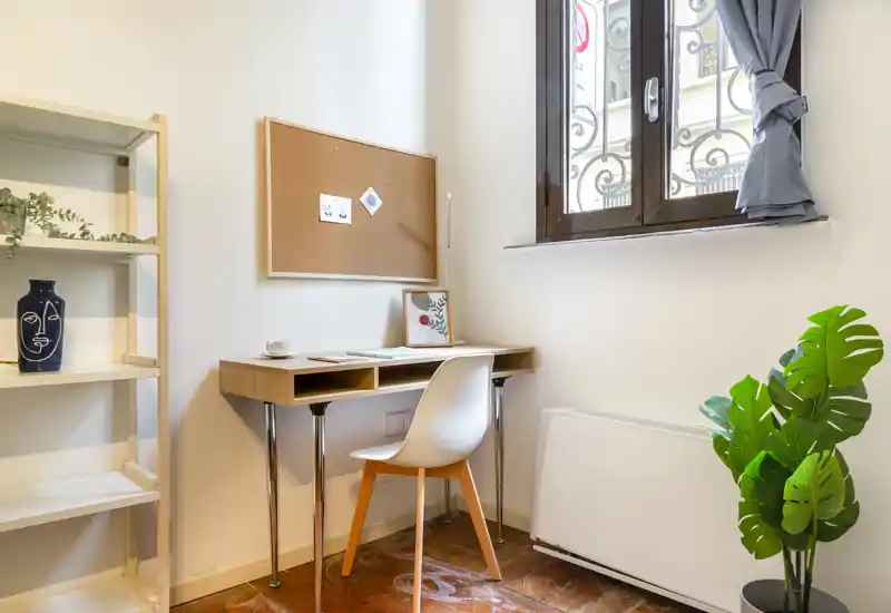 Angolo soggiorno/studio luminoso e compatto con una piccola scrivania in legno, sedia e una bacheca in sughero accanto alla finestra — arredato con piante e scaffali semplici per un angolo studio accogliente.