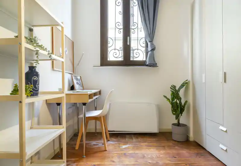 Stanza compatta e luminosa con una scrivania e scaffalatura, pavimento in legno e armadio — perfetta come angolo camera/studio con luce naturale dalla finestra.