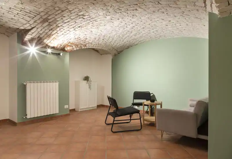 Zona giorno minimal e accogliente con soffitto a volte in mattoni, parete accentuata verde chiaro, pavimento in cotto e sedute semplici (divano e sedie) intorno a un tavolino.