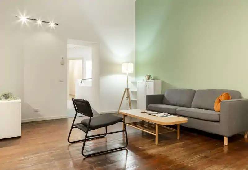 Soggiorno moderno e luminoso con divano grigio, tavolino in legno e poltrona di design su parete verde chiaro; illuminato da una lampada da terra e cuscino arancione per un tocco accogliente.