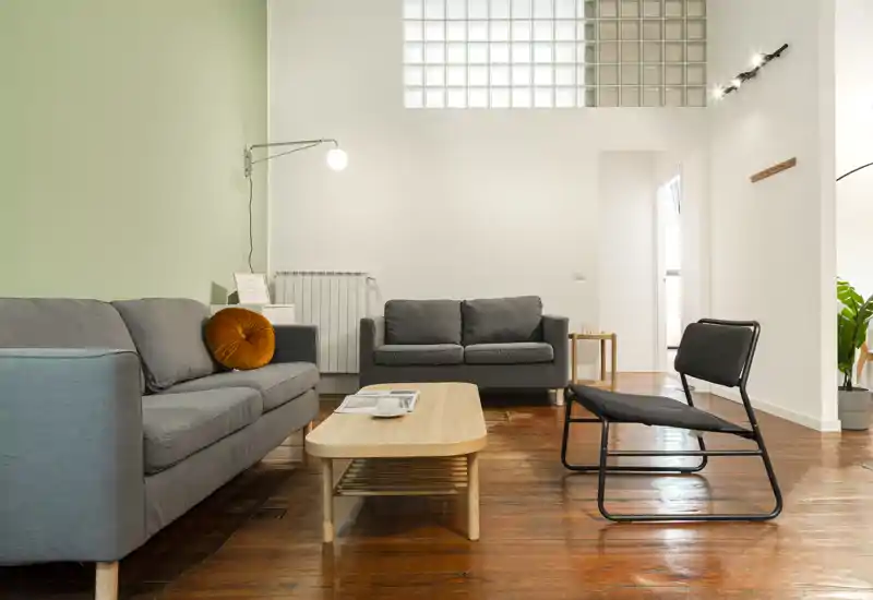 Soggiorno moderno e luminoso con due divani grigi, un tavolino in legno e una poltrona nera minimalista su pavimento in legno caldo — area accogliente e tranquilla ideale per il relax.