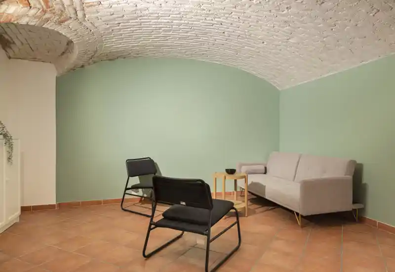 Zona giorno accogliente con divano grigio chiaro, due poltrone nere e un piccolo tavolino in legno sotto una caratteristica volta in mattoni bianchi — pavimento in cotto e pareti verde tenue creano un’atmosfera calma e invitante.