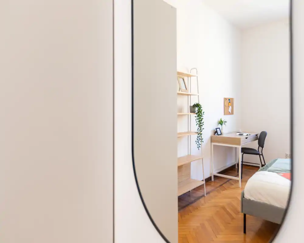 Dormitorio luminoso y minimalista con cama bien hecha, suelo de parquet, un pequeño escritorio y estantería — ideal para una estancia cómoda o como espacio de trabajo.