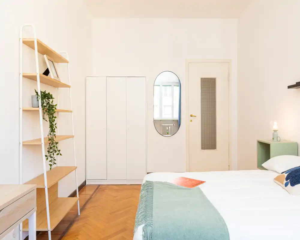 Dormitorio luminoso y ordenado con cama bien hecha, manta verde suave y estantería minimalista. Suelo de parquet cálido, armario y espejo ovalado en la pared que refleja la luz natural.
