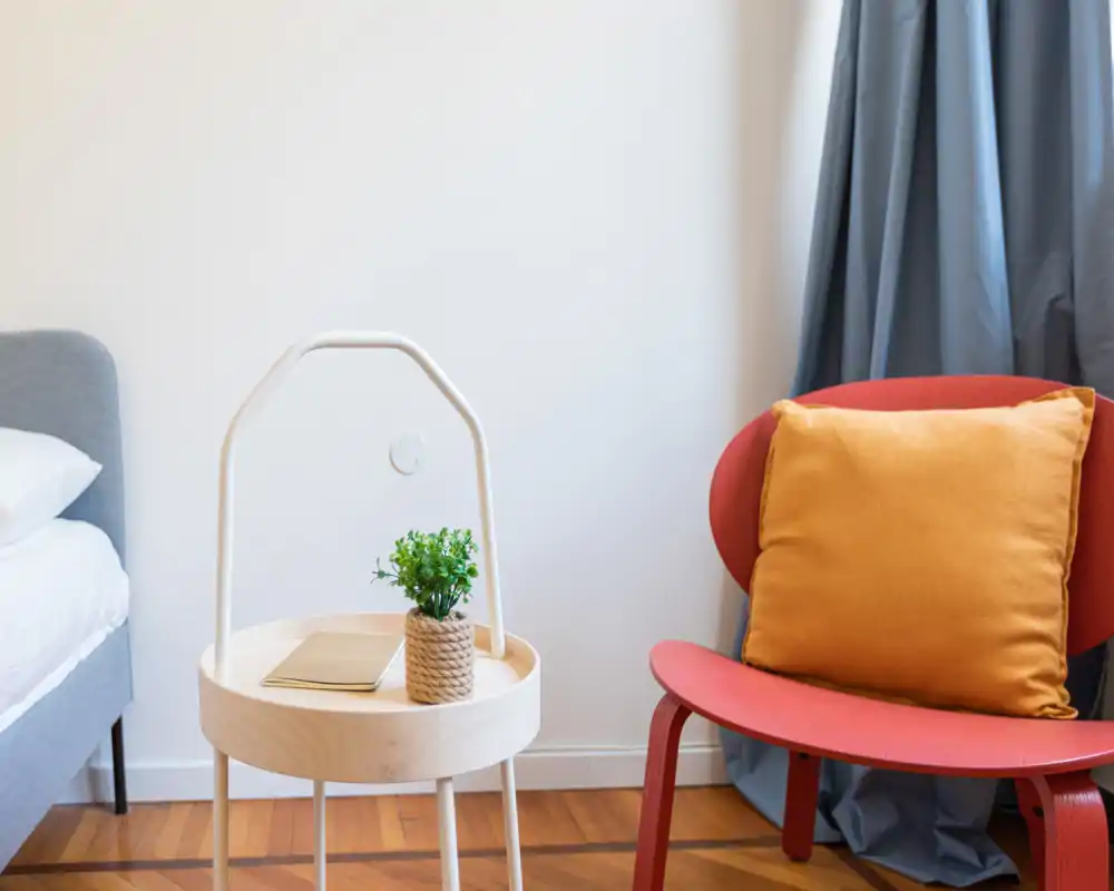 Rincón de dormitorio luminoso y moderno con el borde de la cama visible, una mesita con planta y cuaderno, y una butaca roja con cojín cálido.
