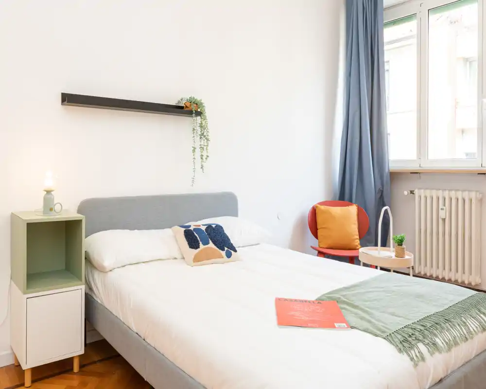 Dormitorio moderno y luminoso con cama doble bien hecha, cojín decorativo, mesita y una ventana que aporta abundante luz natural.