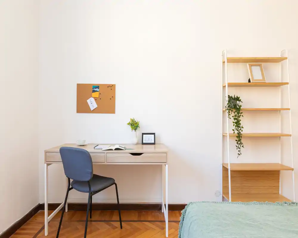Rincón de dormitorio luminoso y minimalista con escritorio de madera clara, silla y parte de la cama visible; estantería sencilla y plantas crean un ambiente limpio y aireado.