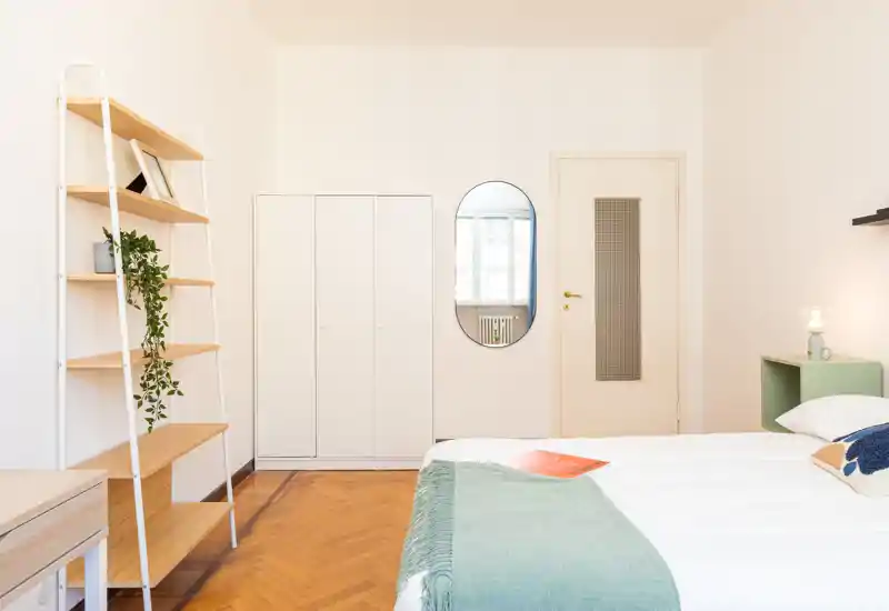 Camera da letto luminosa e minimal con letto ben rifatto, plaid verde chiaro e scaffalatura semplice. Ambiente arioso con luce naturale e pavimento in parquet pulito.